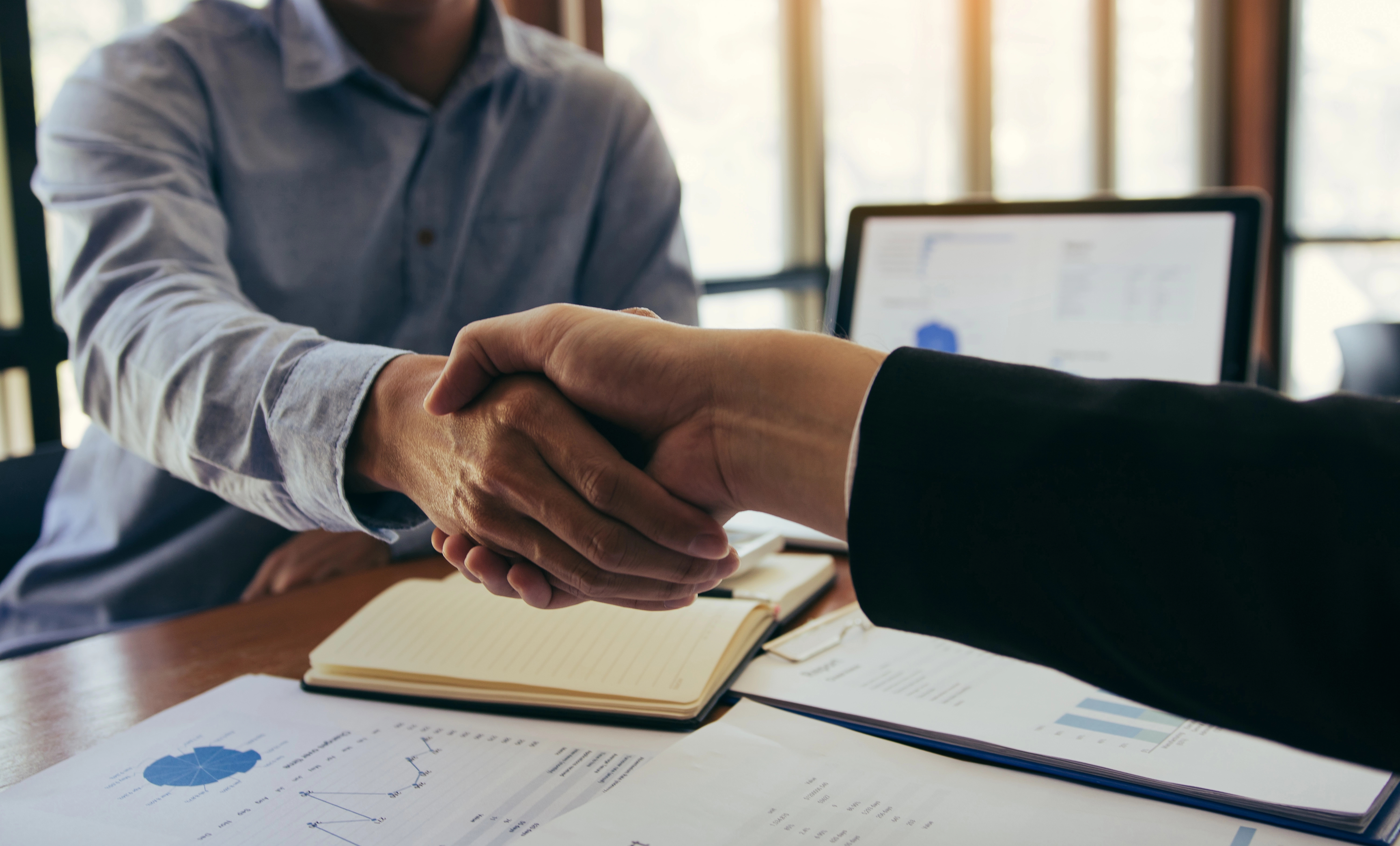 Two people shaking hands, papers on desk, laptop in background, Depositphotos 374370094 Xl